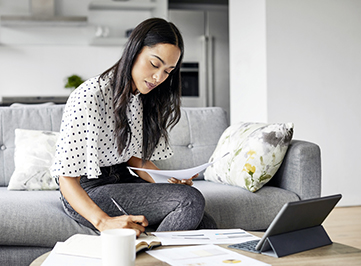 young adult woman sitting on the couch working on paperwork with her ipad